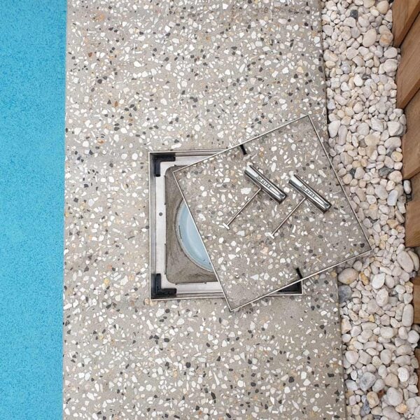 HIDE stainless steel skimmer cover opened in exposed aggregate paving, showing the recessed inlay lid and lifting keys beside the pool.