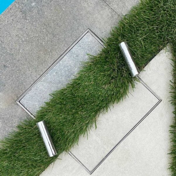 HIDE stainless steel Skimmer Cover set flush within artificial turf and pavers, shown with two lifting keys for easy removal.