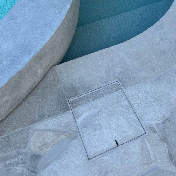 HIDE stainless steel skimmer cover set flush in stone paving beside curved pool steps; joint lines continue through the cover to match the surrounding pattern.
