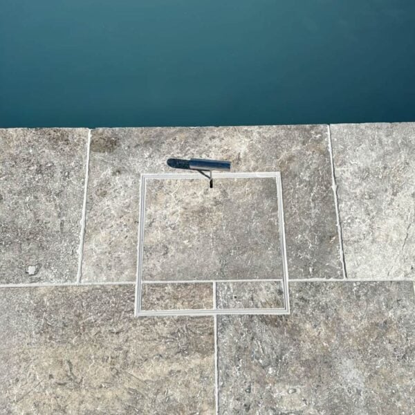 HIDE stainless steel skimmer cover set flush in stone pavers beside the pool; grout lines align through the cover, shown with safety key.