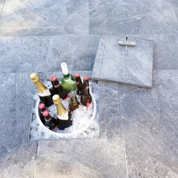 HIDE stainless steel access cover opened in stone pavers, revealing a recessed ice bucket with bottles inside, shown with lifting key.