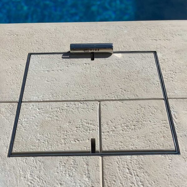 HIDE stainless steel skimmer cover set flush in light stone coping; grout lines continue through the cover, shown with safety lifting key.