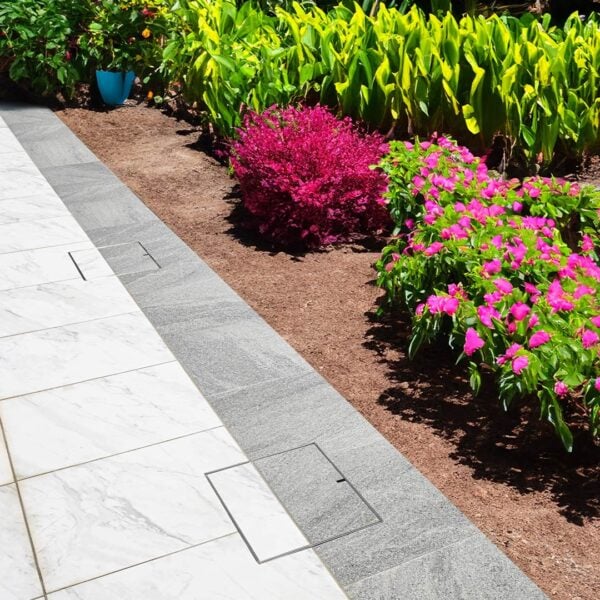 Two stainless steel HIDE Access Cover with dual grey and white tile inlay installed next to bright vibrant garden.
