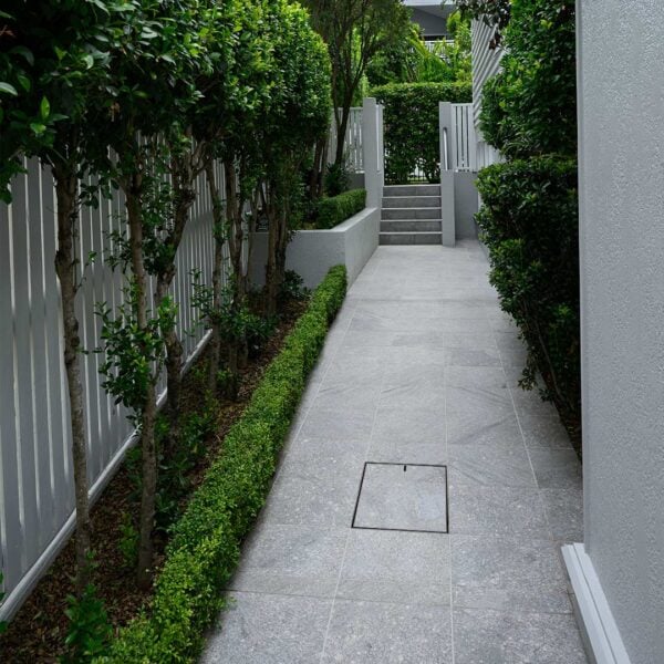 Flush finish HIDE Drain Cover installed in walkway between a house and fence.